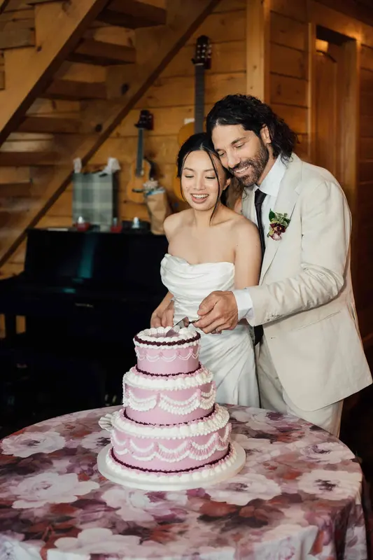 Ji and Noah cutting a tiered pink and white cake by the piano in the great room