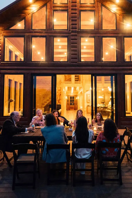 Guests at dinner tables on the deck at dusk in front of the lit glass gable