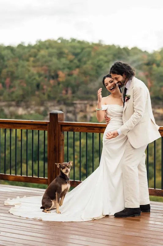 Ji laughing in Noah's arms at the deck railing while a small brown dog sits on her train, with the cliffs and fall forest behind