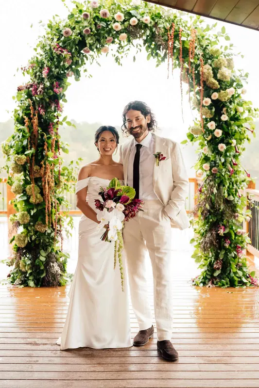 Ji and Noah standing under a floral arch on the deck, holding a bouquet of red and white flowers