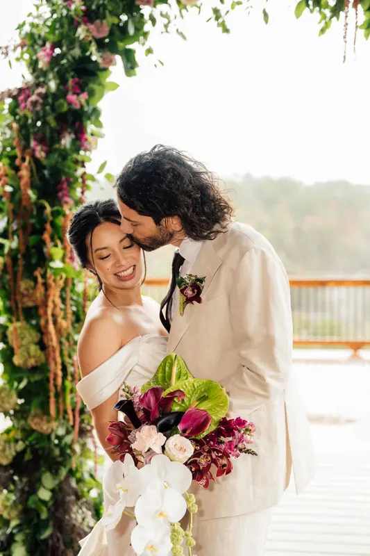 Noah kissing Ji's forehead as she holds her bouquet, with the gorge in soft focus behind them