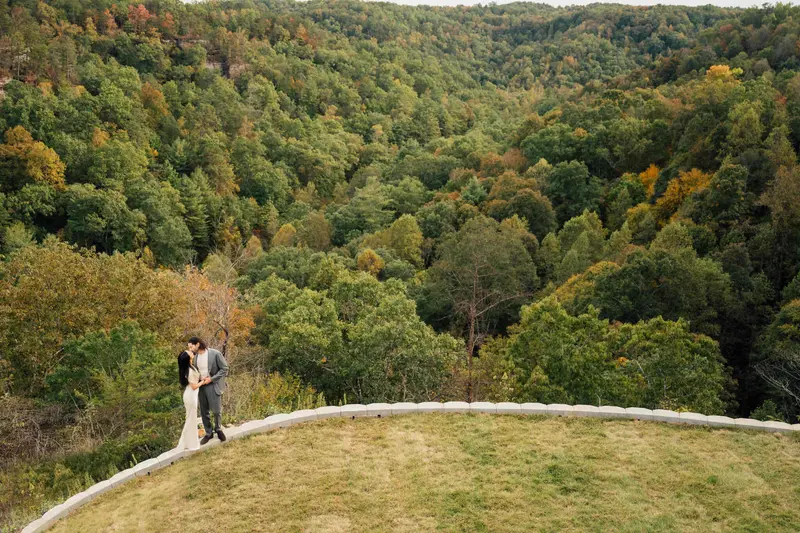 The couple small on the lawn at the cliff's edge with a wide forested gorge spread out below