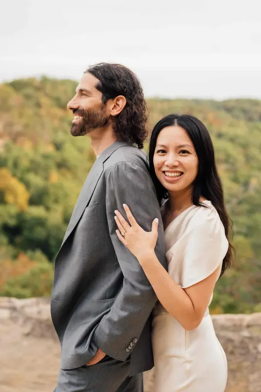 The couple smiling on a sandstone overlook with fall-tinged forest behind them
