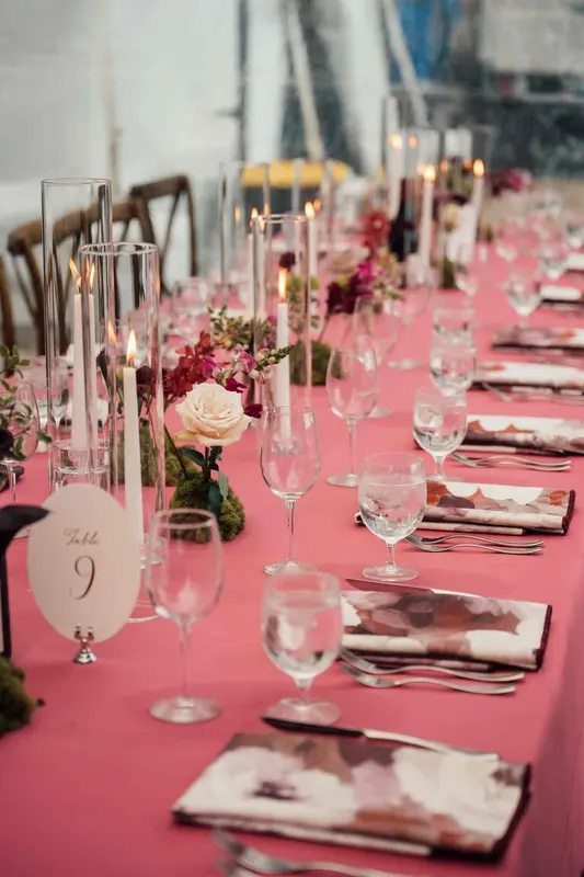 A long reception table with a pink cloth, taper candles, glassware and a table number card