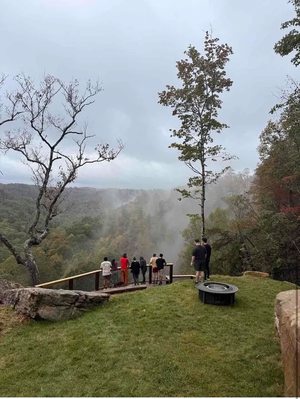 Guests at the cliff-edge overlook railing watching mist rise out of the gorge, with a fire pit on the lawn
