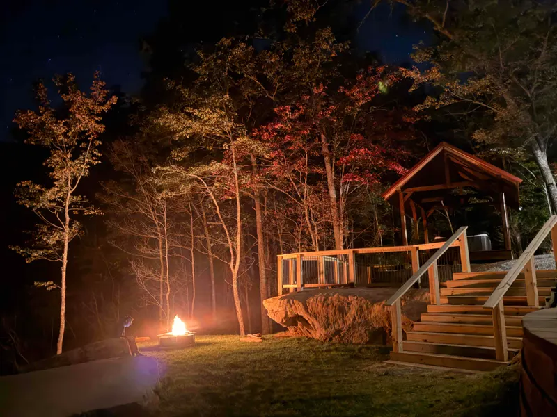 A lit fire pit on the lawn at night beside a wooden pavilion on a sandstone boulder, with lit fall trees behind