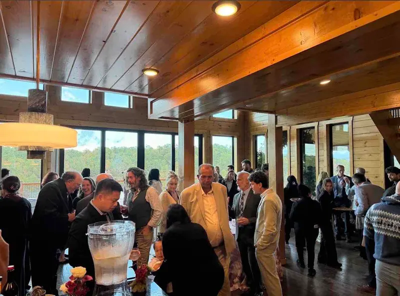 A crowd of guests mingling in the great room, with windows to the forest behind them