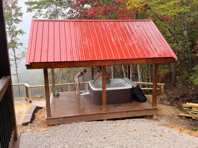 Guests getting into the hot tub under a covered pavilion with a red metal roof, with fall trees and mist over the gorge behind the deck railing