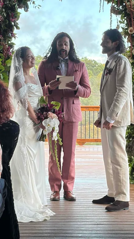 A bride and groom exchanging vows on the deck under a floral arch, with the officiant reading and the forest behind